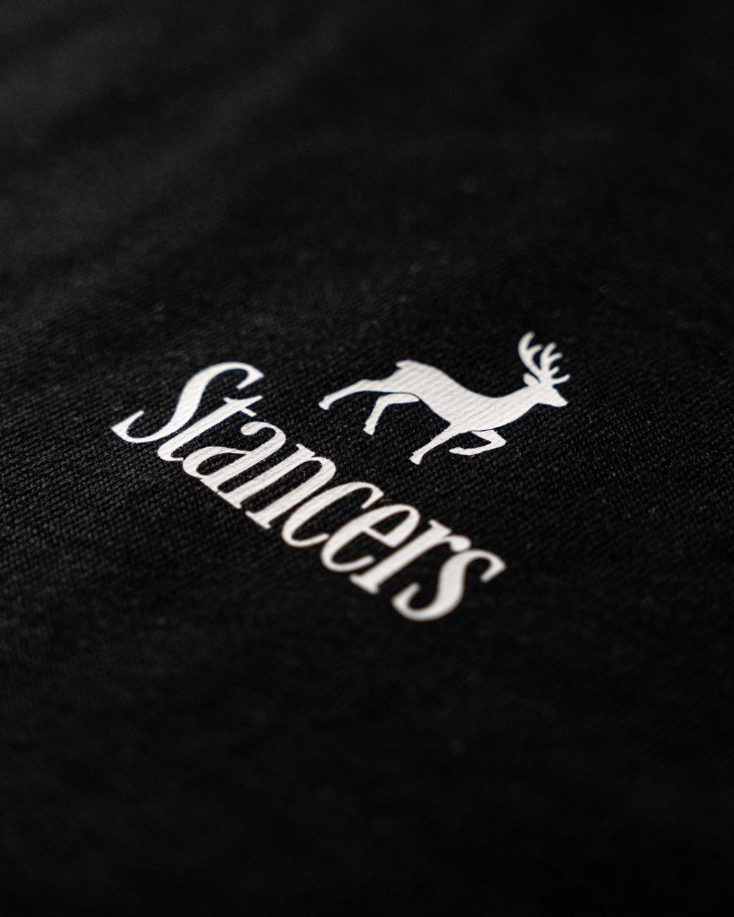 Stancers Logo Branding – Minimalist Streetwear Detail Minimalistisches weißes „Stancers“ Logo mit Hirsch-Symbol auf der Brust eines schwarzen Premium-T-Shirts.